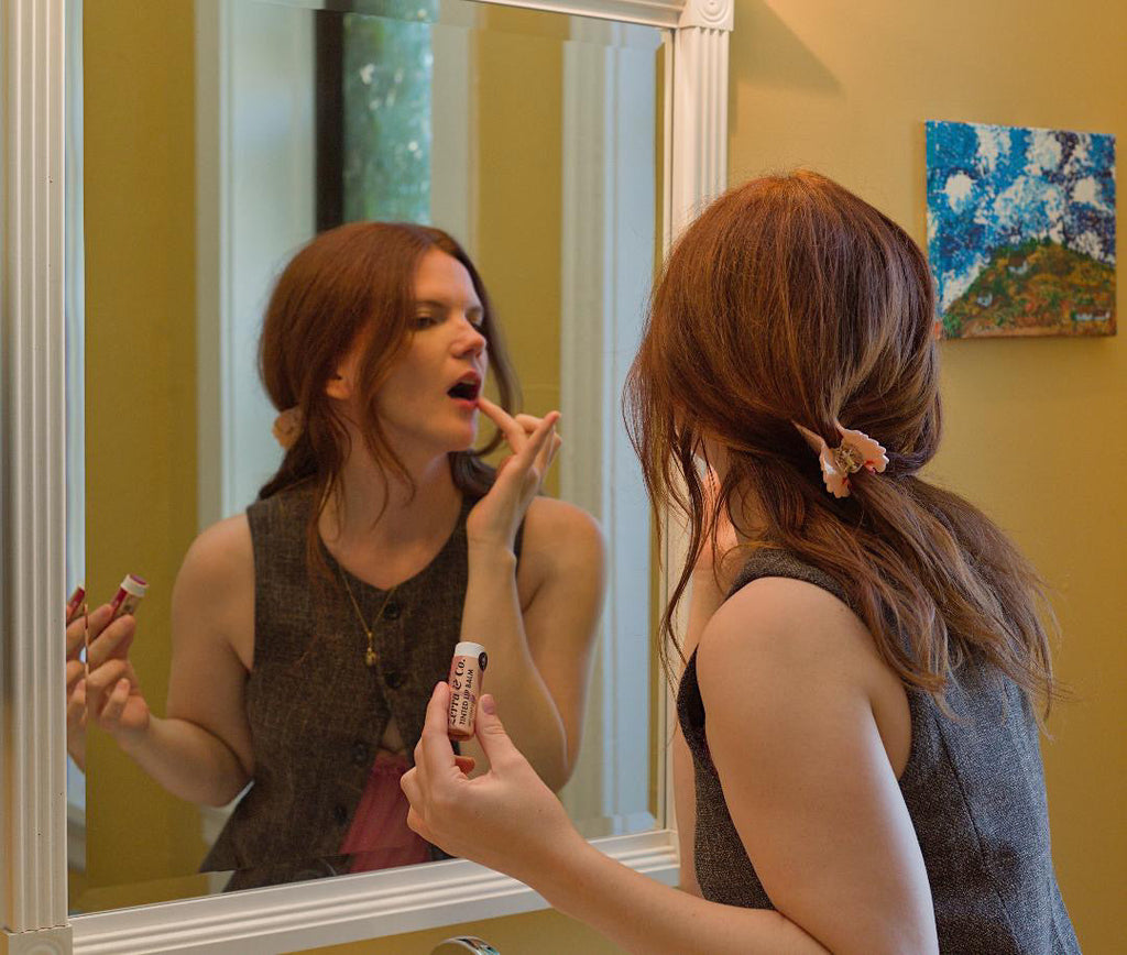 A young woman does her makeup in the mirror and shows off a plastic-free tube of lip balm.