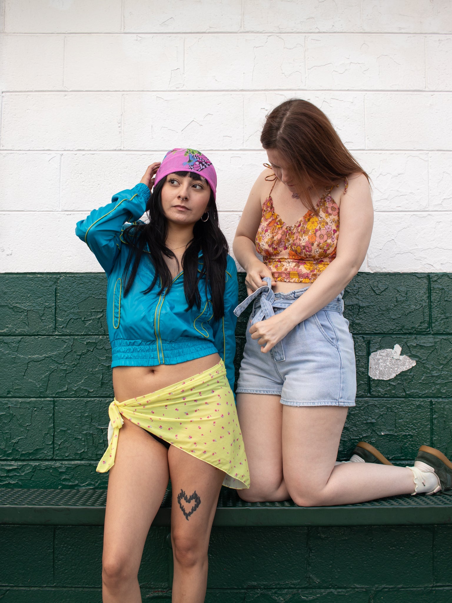 Two young women posing together on a dark green bench against a white brick wall. The woman on the left wears a bright blue zip-up jacket, a butterfly patterned head scarf, and a yellow patterned sarong tied around her thigh.