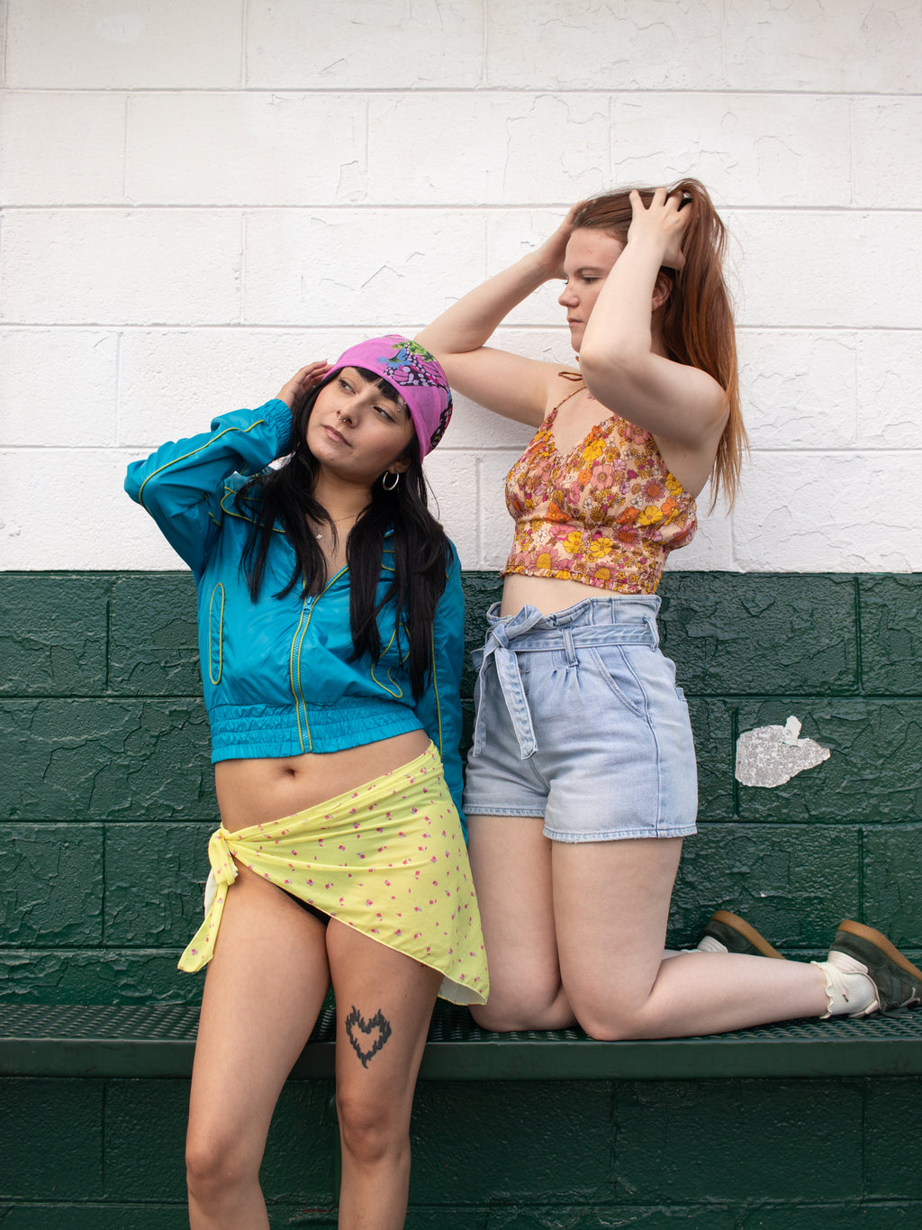Two young women posing together on a dark green bench against a white brick wall. The woman on the left wears a bright blue cropped zip-up jacket, a pink patterned head scarf, and a yellow sarong.