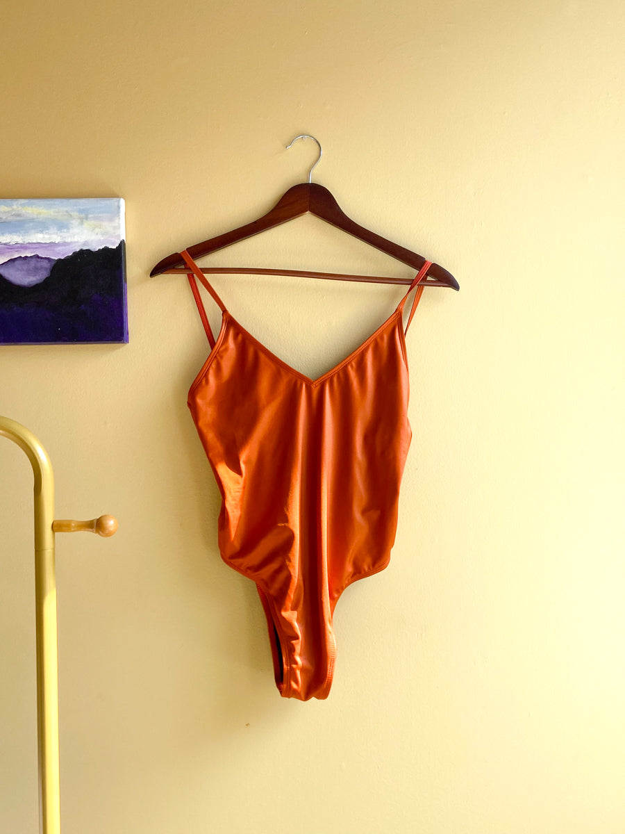 A rust-colored bathing suit with spaghetti straps hanging on a dark wooden hanger against a beige wall.